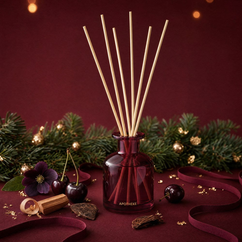 Burgundy Bitter Cherry diffuser surrounded by evergreen branches, dark cherries, purple flowers, wood pieces, and curled burgundy ribbon, with warm lights against a deep red backdrop.