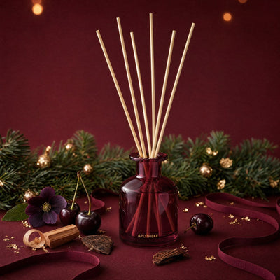 Burgundy Bitter Cherry diffuser surrounded by evergreen branches, dark cherries, purple flowers, wood pieces, and curled burgundy ribbon, with warm lights against a deep red backdrop.