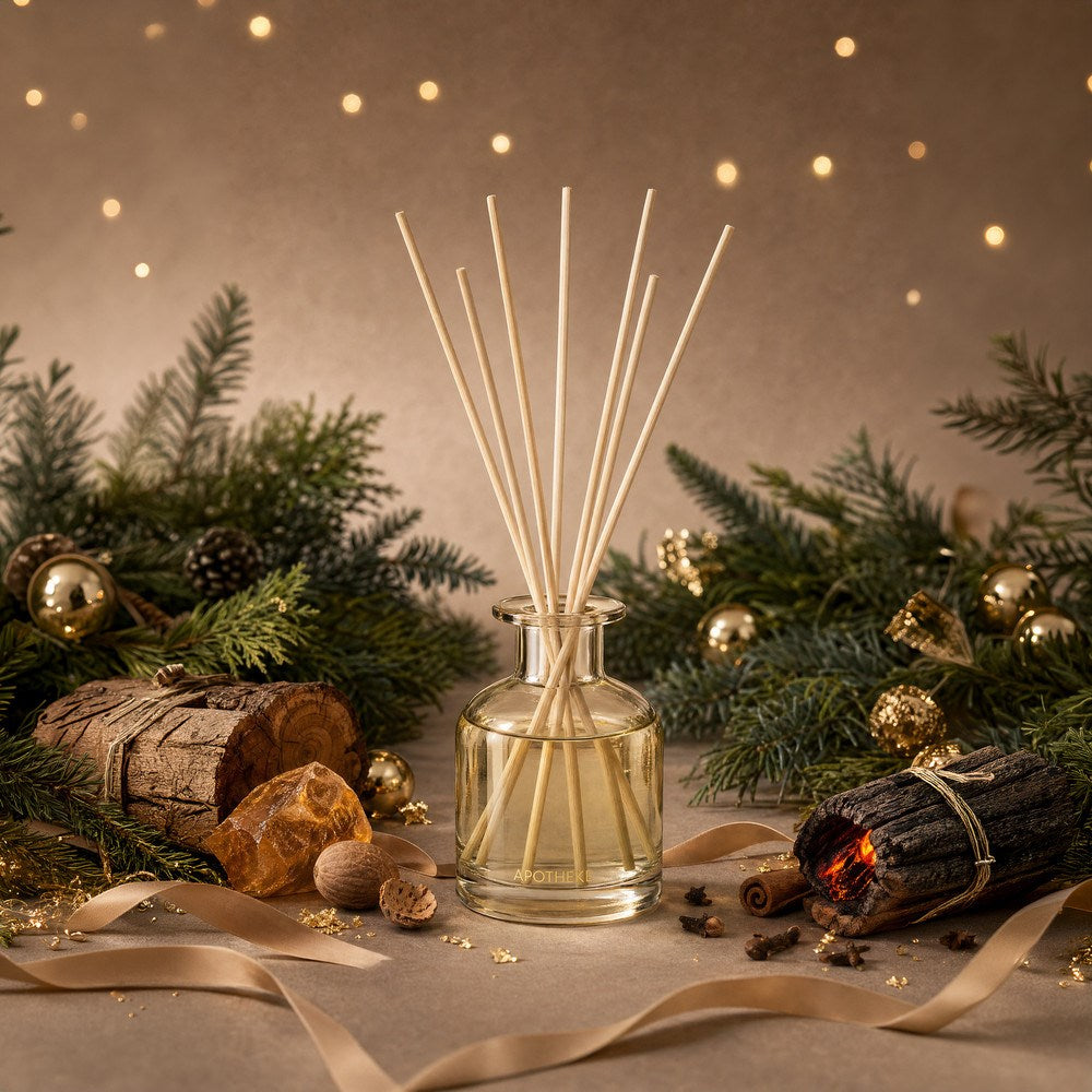 Firewood Mini Reed Diffuser surrounded by evergreen branches, gold ornaments, bundled wood, spices, and amber-colored pieces, with gold ribbon across the tabletop and warm lights behind.