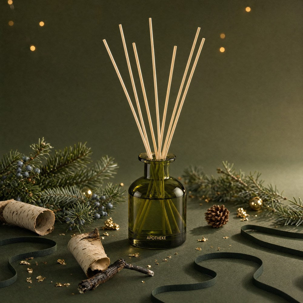 Green Juniper Birch diffuser surrounded by evergreen branches, blue berries, white birch logs, pinecones, and curled dark green ribbon, with scattered gold accents and warm lights behind.