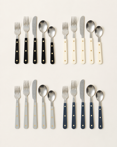 Four five-piece Heritage Cutlery place settings in black, ivory, pale blue-gray, and navy, each with two forks, a knife, a tablespoon, and a teaspoon featuring polished metal heads and triple-riveted handles.