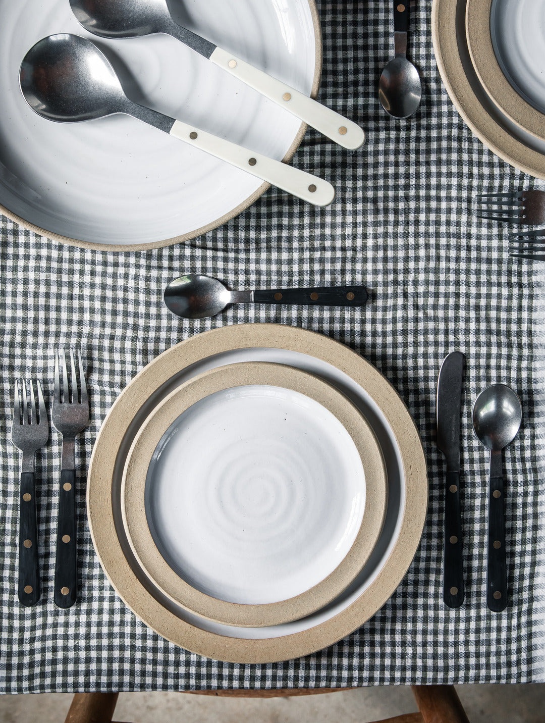 Overhead view of black-handled Heritage Cutlery set around layered neutral and white plates on a black-and-white gingham tablecloth, with ivory serving utensils nearby.