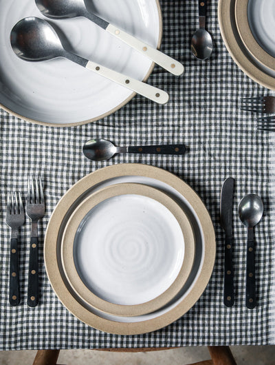 Overhead view of black-handled Heritage Cutlery set around layered neutral and white plates on a black-and-white gingham tablecloth, with ivory serving utensils nearby.