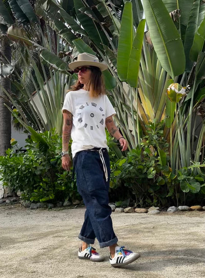 Model walking outdoors in the light gray Service Tee, navy drawstring pants, colorful sneakers, and a beige hat.