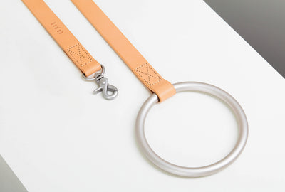Close-up of the peach leather-like Lumi Leash with a brushed-silver swivel clasp, perforated stitched details, and oversized circular metal handle.
