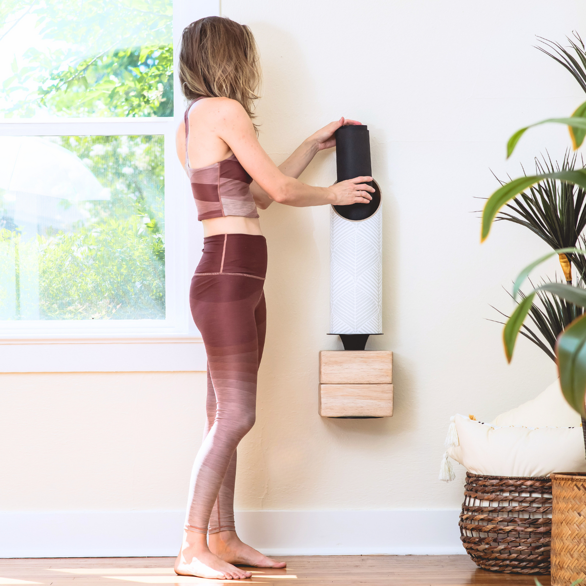 Woman in mauve activewear placing a rolled black yoga mat into a wall-mounted white Sattva tube.