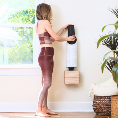 Woman in mauve activewear placing a rolled black yoga mat into a wall-mounted white Sattva tube.