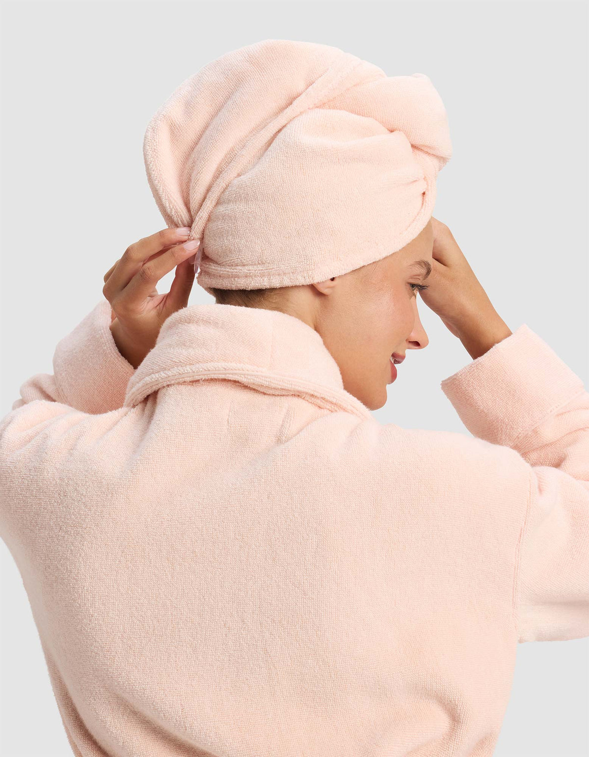 Rear view of the pale peach hair towel wrap, with the model raising both hands to adjust it while wearing a matching robe.