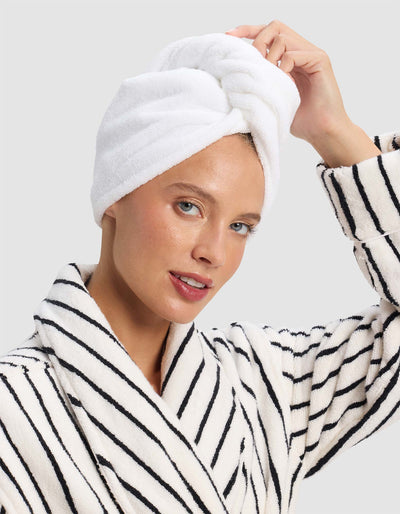 Model adjusting a white Luxe Hair Towel twisted into a wrap on top of her head, wearing a white robe with narrow black stripes.