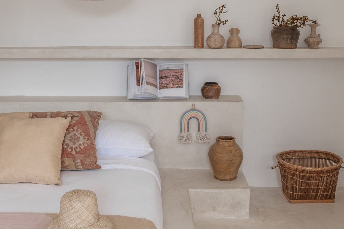 Village Thrive Mia Macrame Rainbow, pastel multicolor cotton, hanging above a bed with pillows and a woven basket.