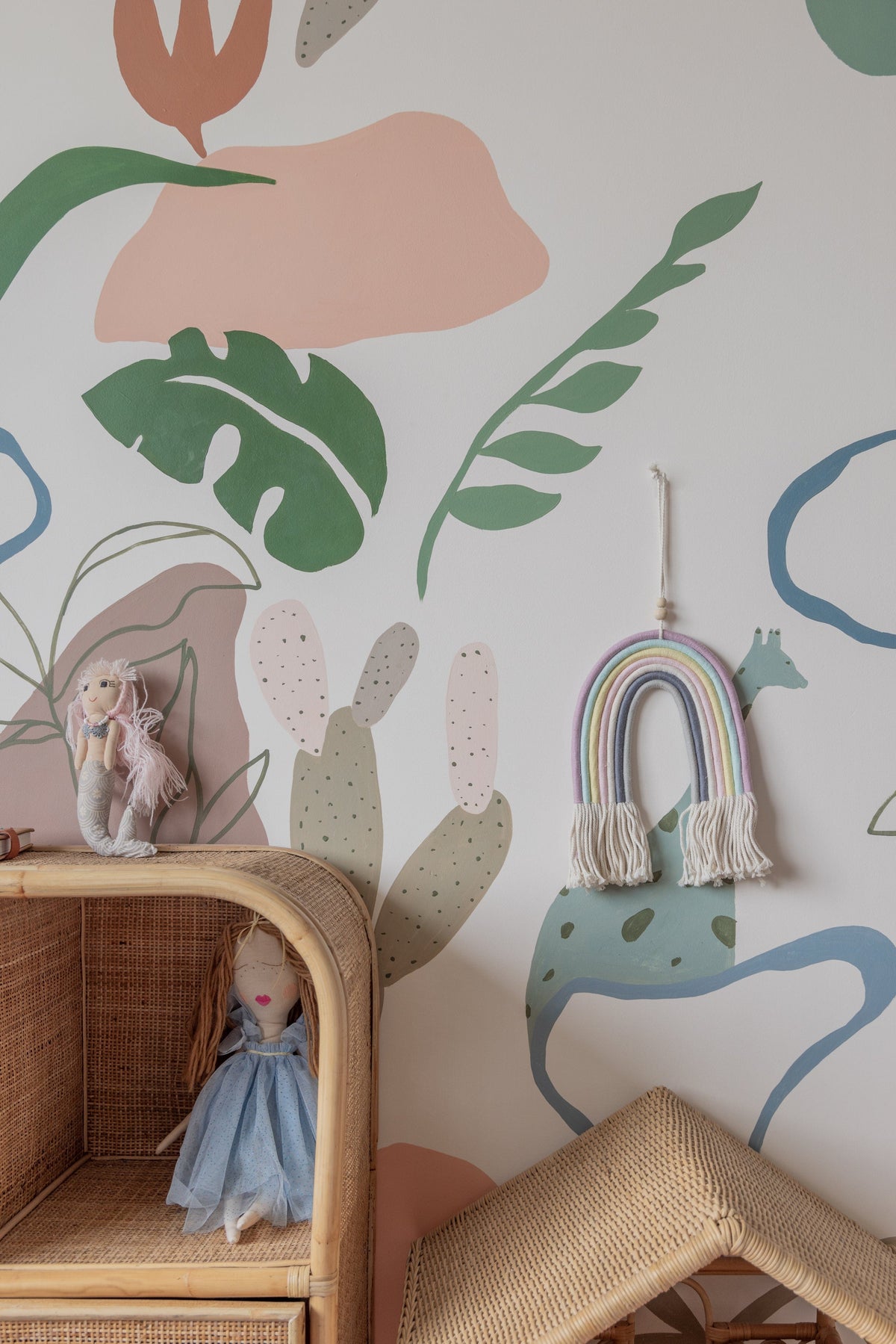 Village Thrive Mia Macrame Rainbow, terracotta and cream cotton, hanging in a nursery beside a floral wall mural and rattan chair.