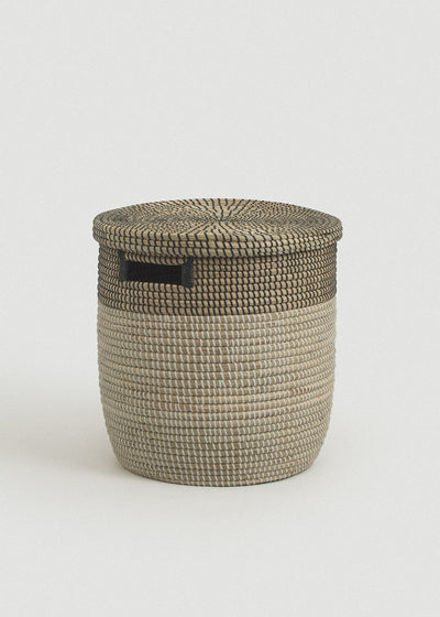 Two-tone woven seagrass storage basket with a natural body, black upper band, flat lid, and rectangular black handle.