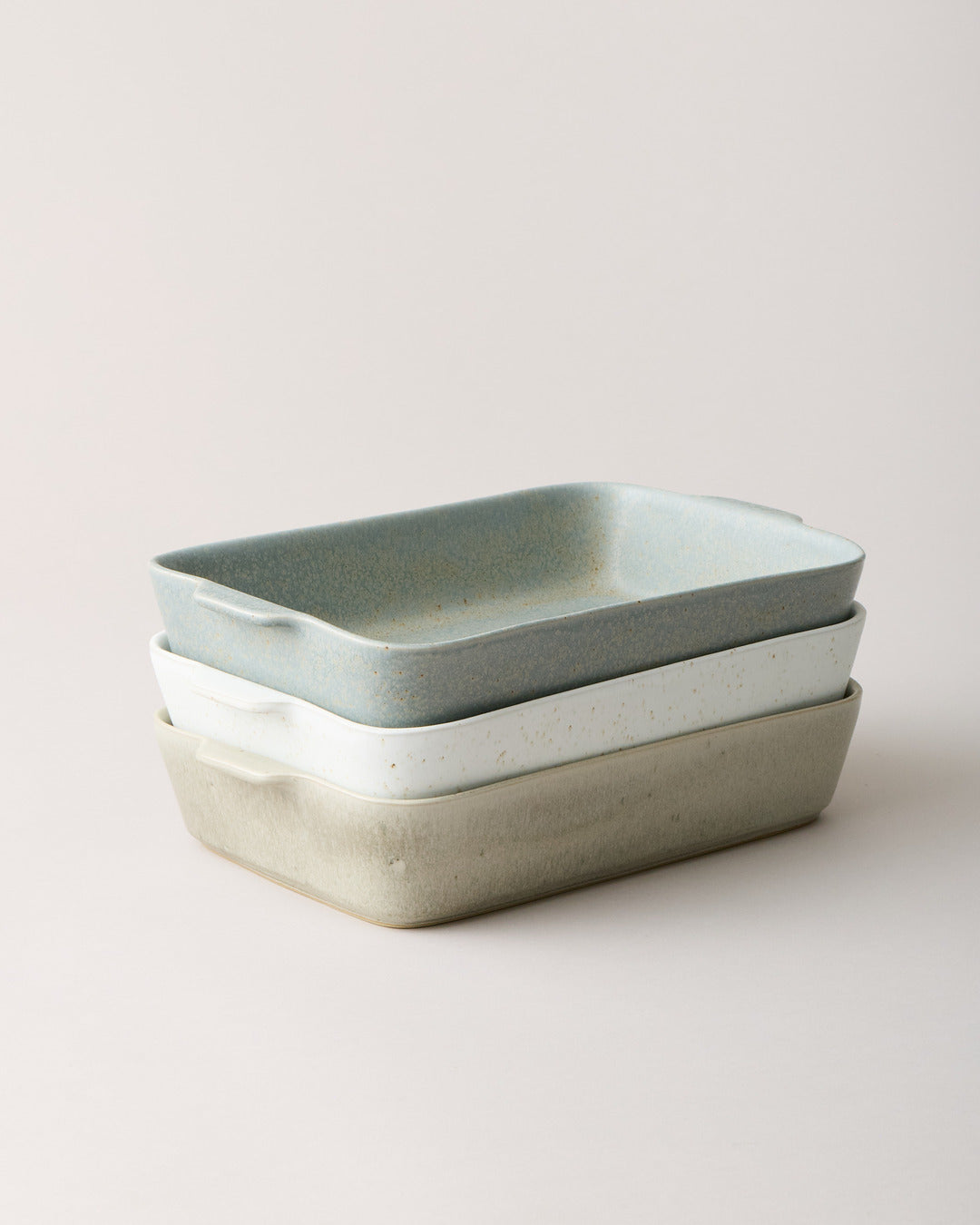 Three rectangular stoneware bakers stacked in soft blue-gray, speckled white, and warm stone glazes, each with rounded corners, gently flared sides, and small integrated side handles.