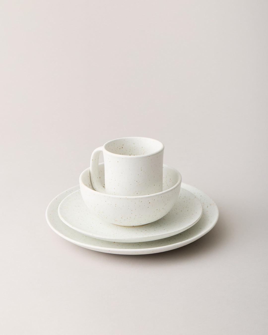 White speckled stoneware mug nested inside a bowl and two plates against a softly lit neutral backdrop.