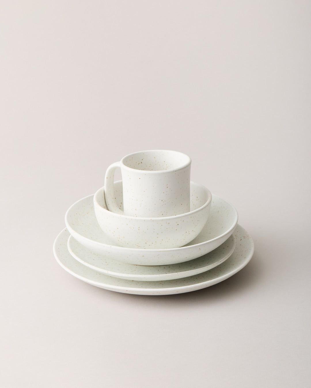 White speckled stoneware place setting with a mug, bowl, and three graduated plates stacked against a pale neutral background.
