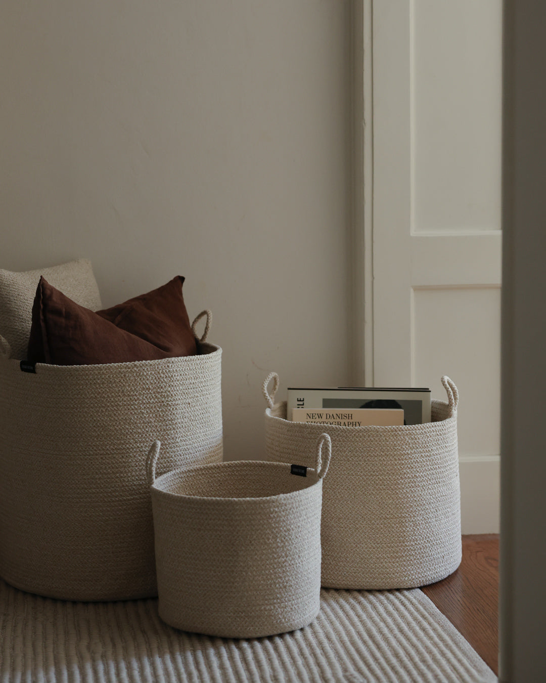 Three beige basket sizes organizing pillows, books, and small objects in a neutral room.