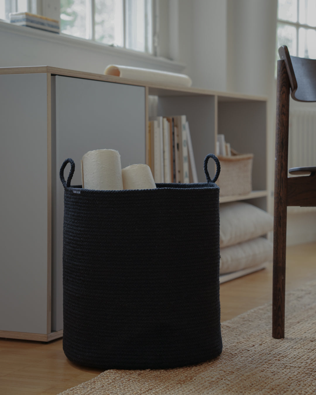 Large black rope basket holding rolled textiles beside a shelving unit in a modern room.