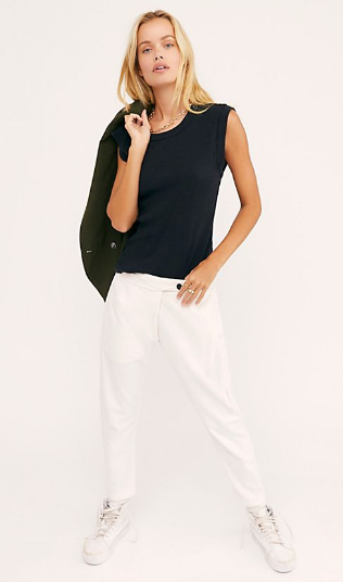 Model wearing white Souk pants with a black sleeveless top, white sneakers, and a black jacket over one shoulder.