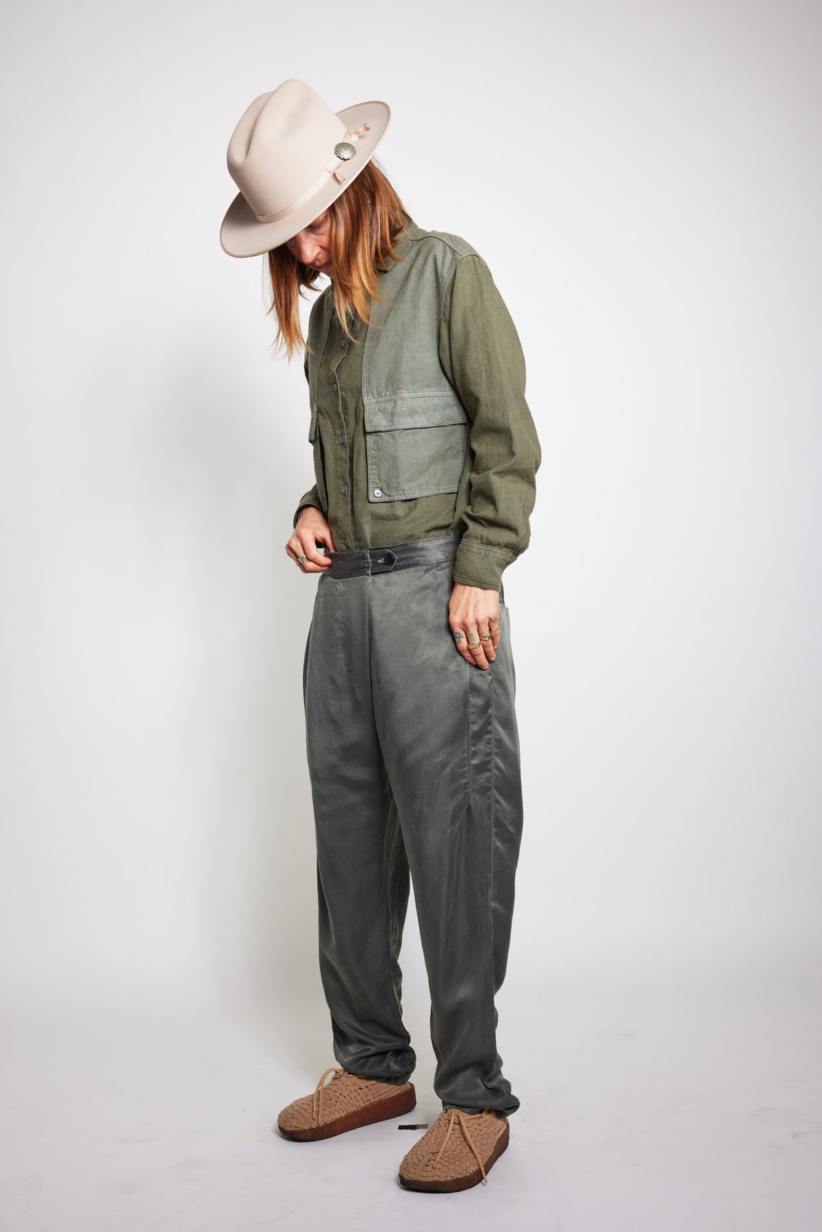 Model looking down while wearing charcoal Souk pants, an olive jacket, and a beige hat.