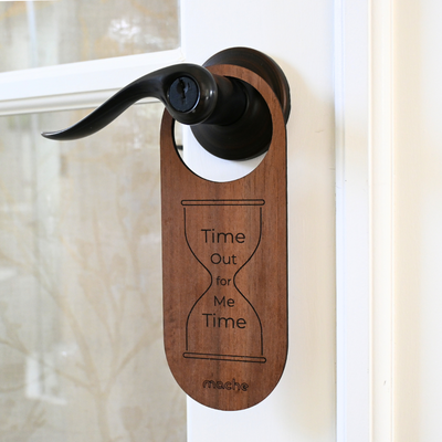 Dark walnut-colored wood door hanger on a black lever handle, featuring the same engraved hourglass design, self-care message, and small Mache logo.