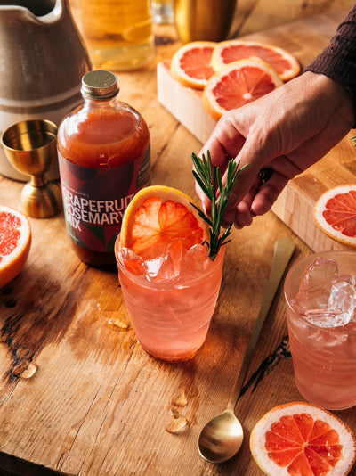 Hand garnishing a pink grapefruit cocktail with rosemary in a clear ridged glass, surrounded by sliced grapefruit and drink-making ingredients.