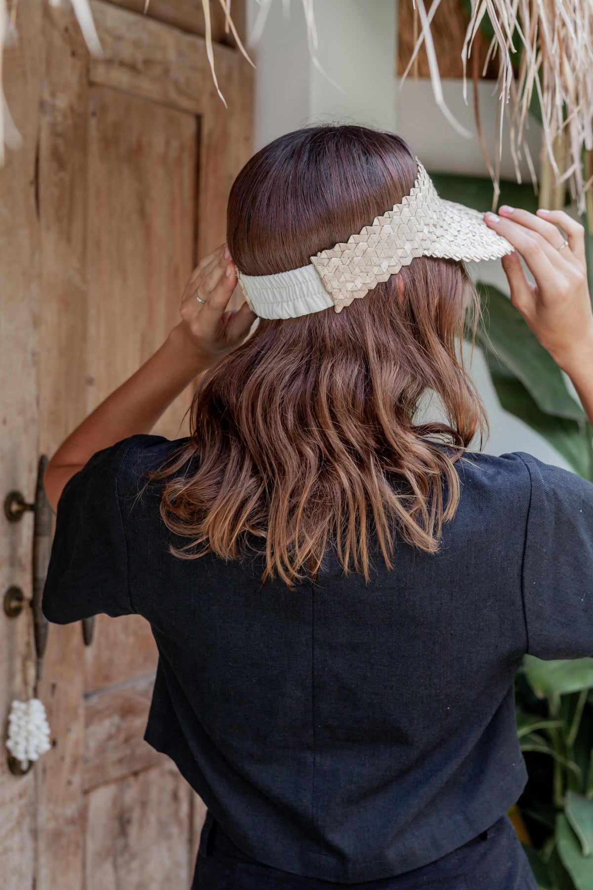 Village Thrive Rattan Sun Visor, blonde palm leaf, worn by a model from behind adjusting the visor.