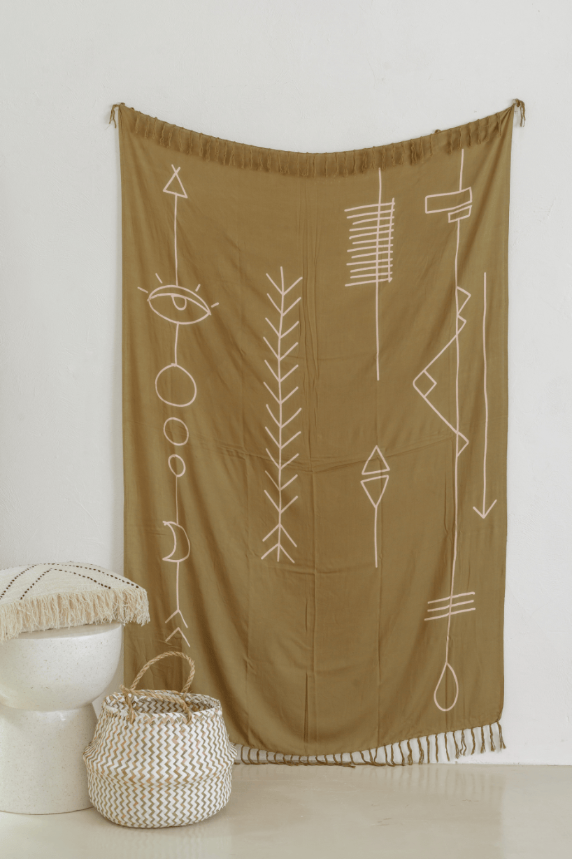 Village Thrive Abstract Tapestry, olive gold rayon, hanging on a wall with a woven basket and ceramic stool below.