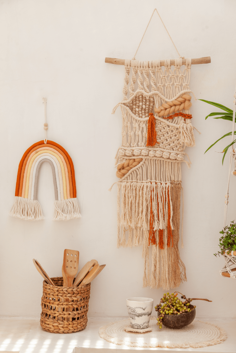Village Thrive Mia Macrame Rainbow, terracotta cotton, hanging beside a larger macrame wall hanging with a potted plant.
