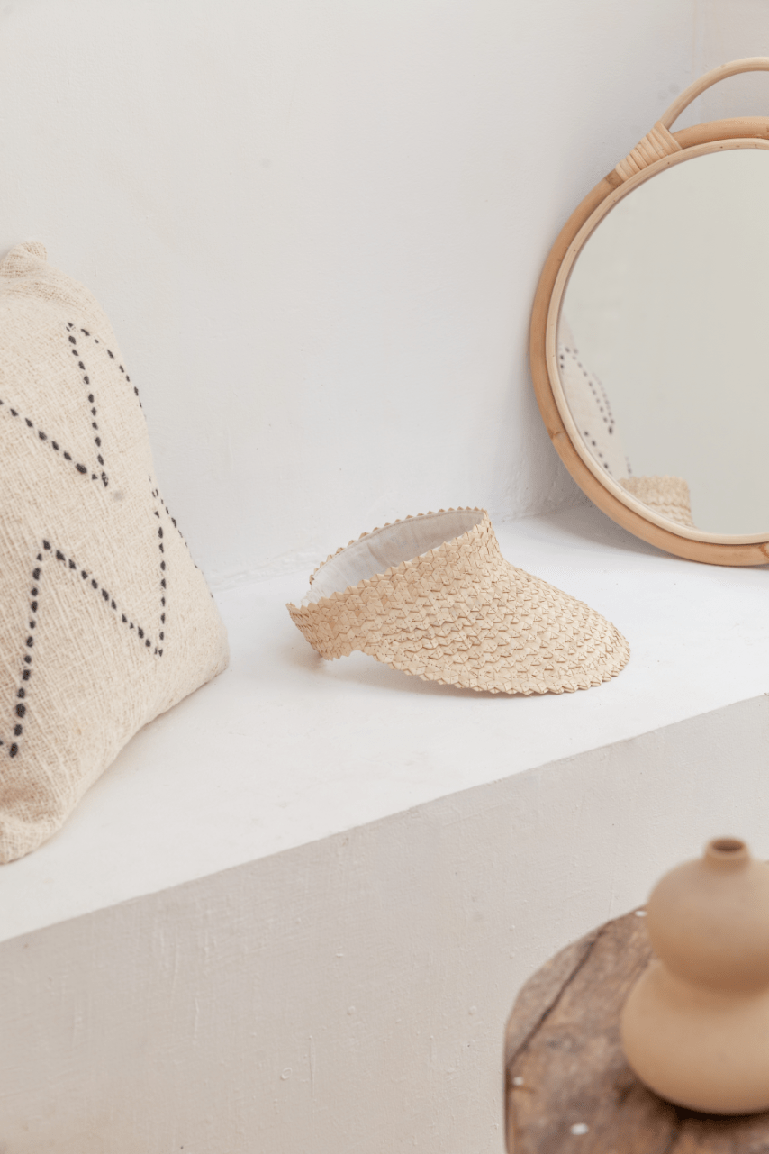 Village Thrive Rattan Sun Visor, blonde and black palm leaf, two visors displayed on a shelf beside a round mirror.