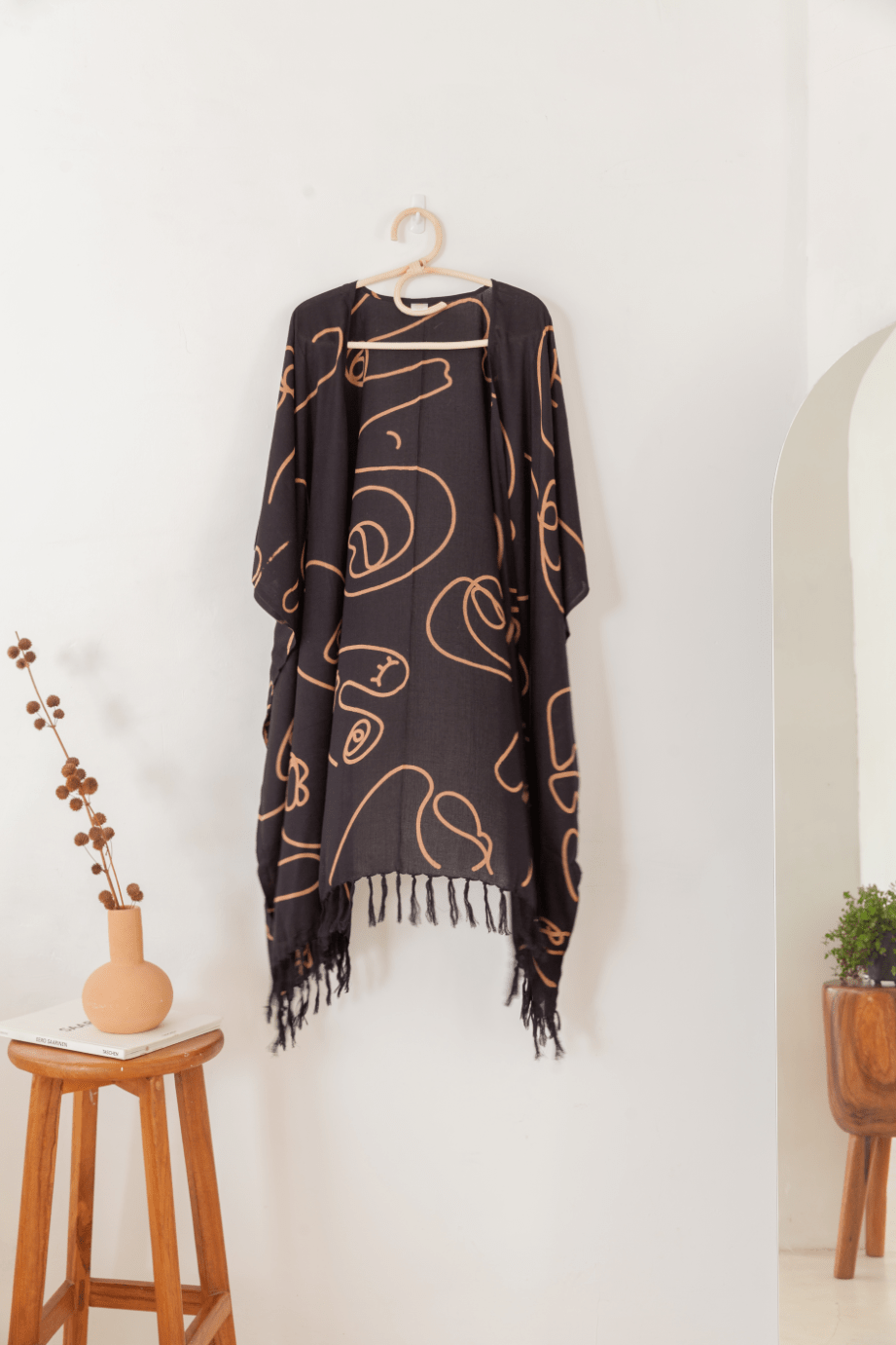 Village Thrive Faces Kimono, black rayon with tan line print, hanging on a wooden hanger against a wall.