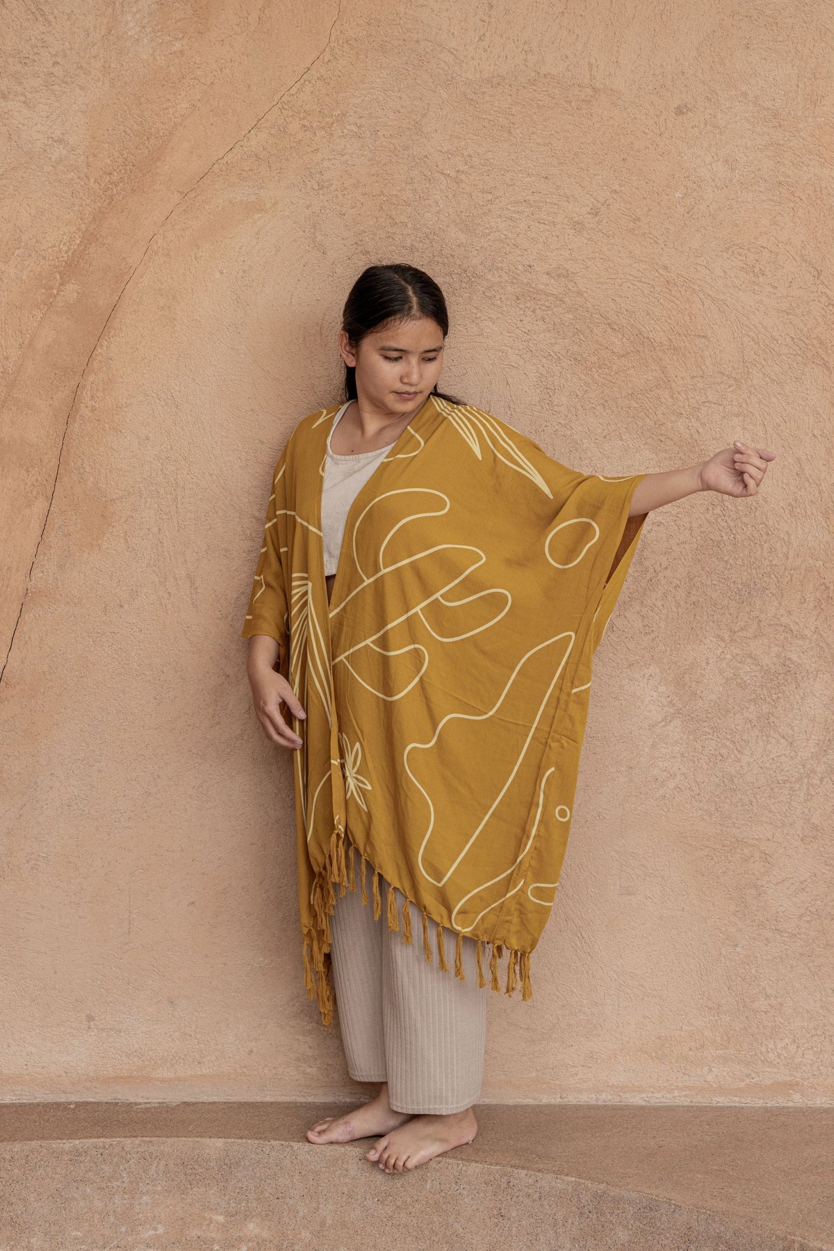 Village Thrive Desert Kimono, mustard gold rayon, worn by a model standing against a stucco wall.