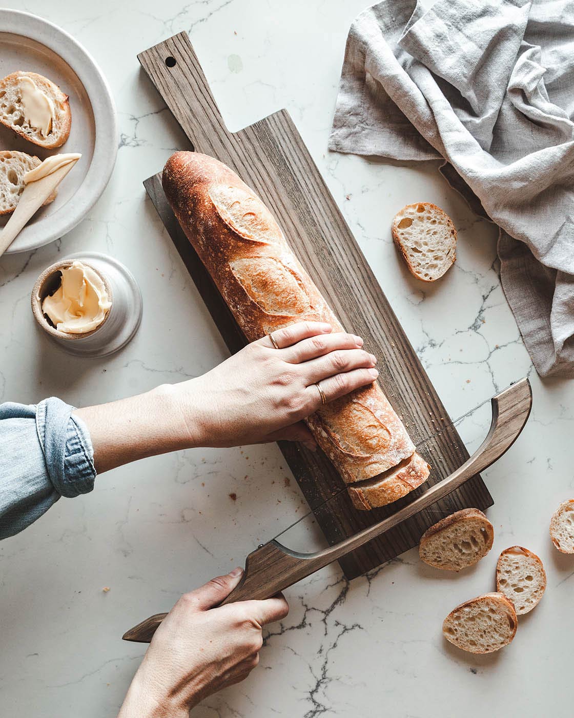 Overhead view of hands slicing a crusty baguette with a wooden bread bow on a narrow cutting board, surrounded by bread slices and gray linen.