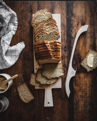 White bread bow beside a sliced seeded loaf on a rustic dark wood table, with bread arranged on a handled cutting board.
