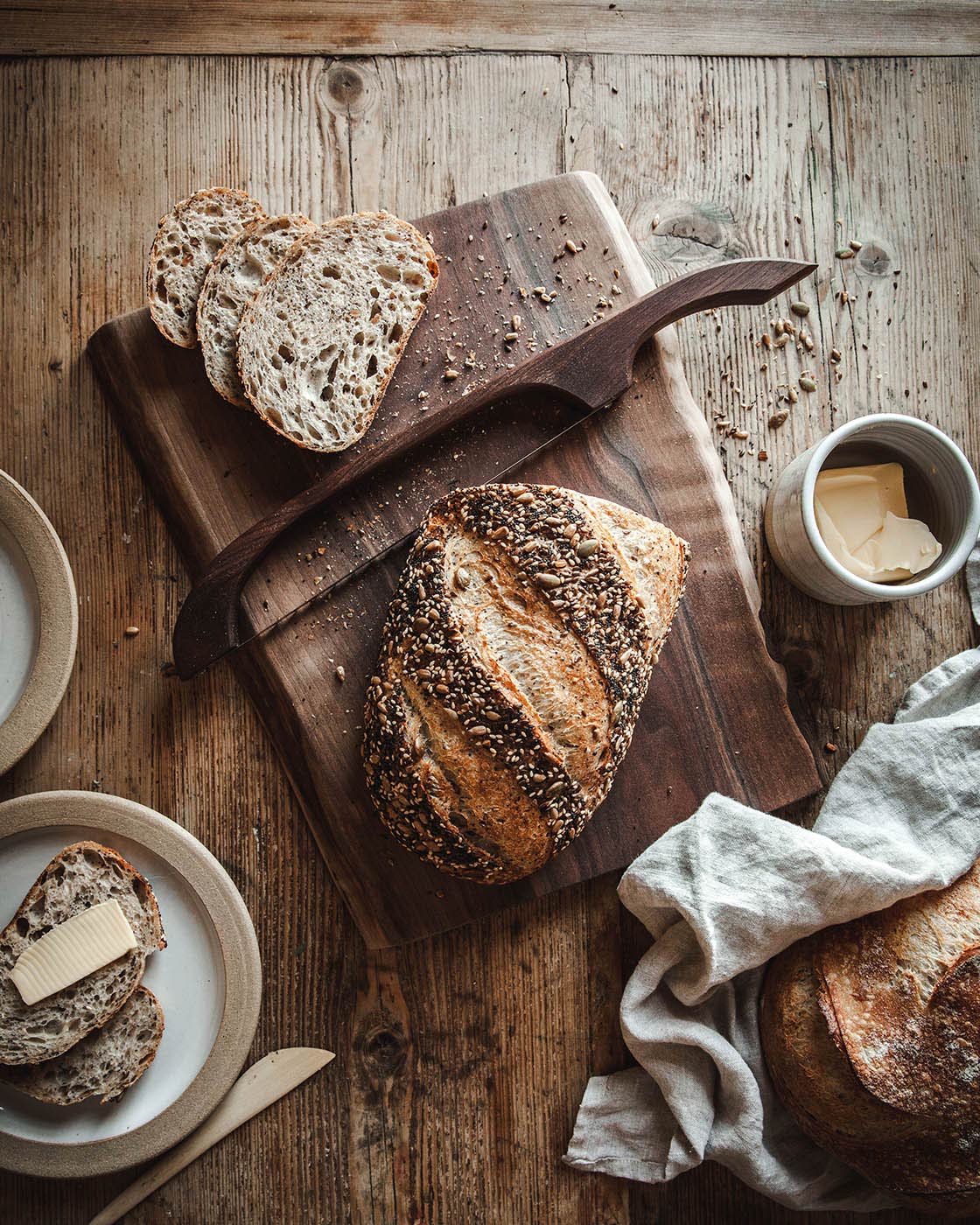 Overhead rustic baking scene with a dark wood bread bow, round artisan loaves, sliced seeded bread, a ceramic cup, and rumpled linen.