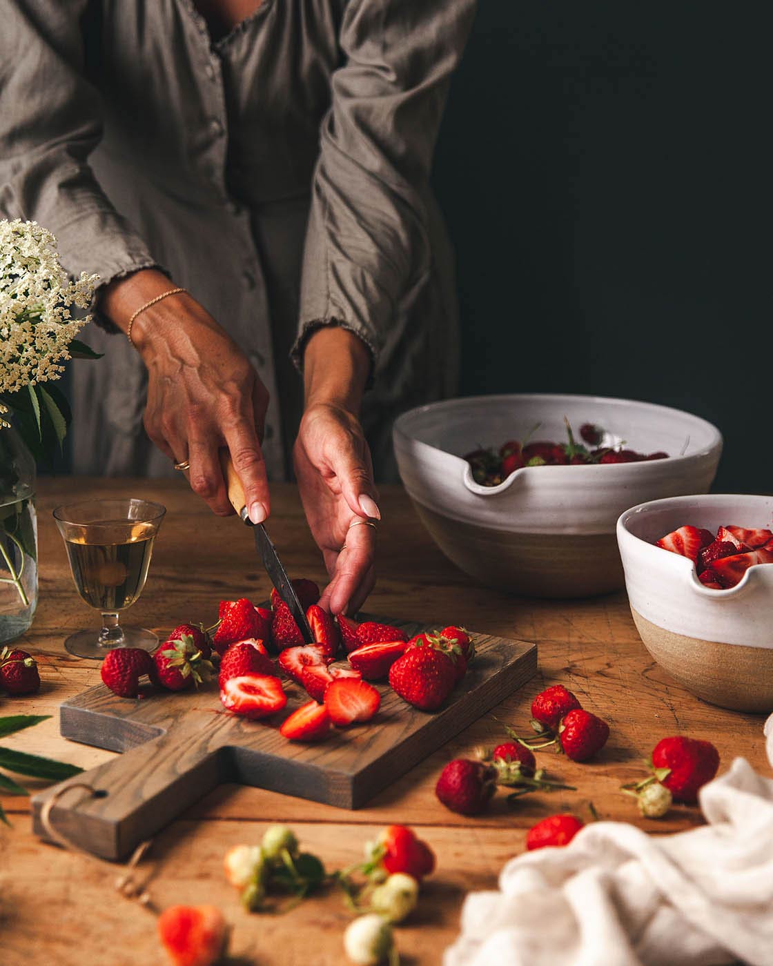 Hands slicing fresh strawberries on a dark wood Pantry Cutting Board surrounded by berries, small bowls, and a glass of amber-colored drink.
