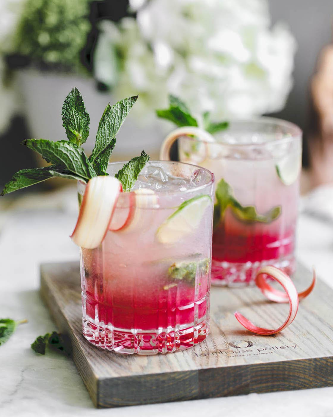 Two pink iced drinks garnished with apple slices and mint, served on a dark wood Pantry Cutting Board used as a tray.