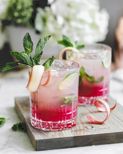 Two pink iced drinks garnished with apple slices and mint, served on a dark wood Pantry Cutting Board used as a tray.