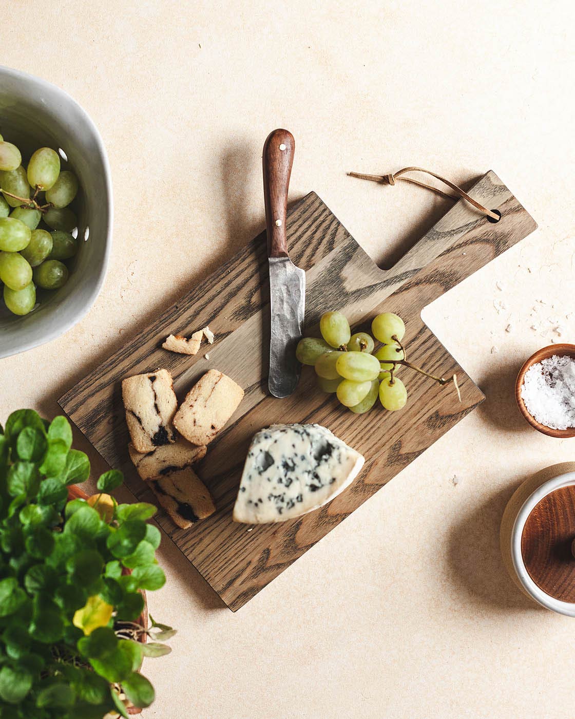 Overhead view of a dark wood Pantry Cutting Board used as a cheese board with blue cheese, sliced bread, green grapes, and a cheese knife.