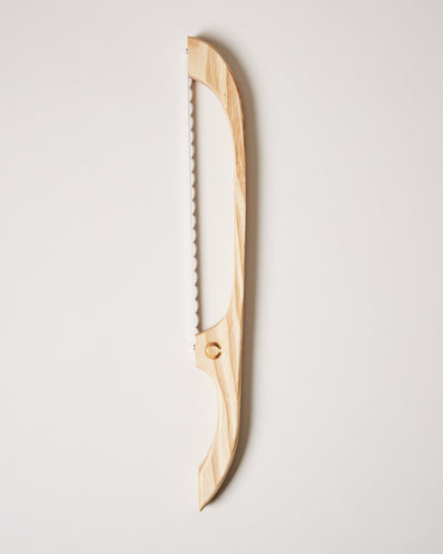 Pale honey-colored wooden bread bow with a serrated blade and sculpted curved handle displayed vertically.