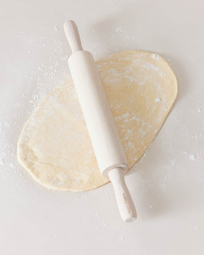 Whitewashed ash rolling pin resting across an oval of flour-dusted dough on a pale work surface.