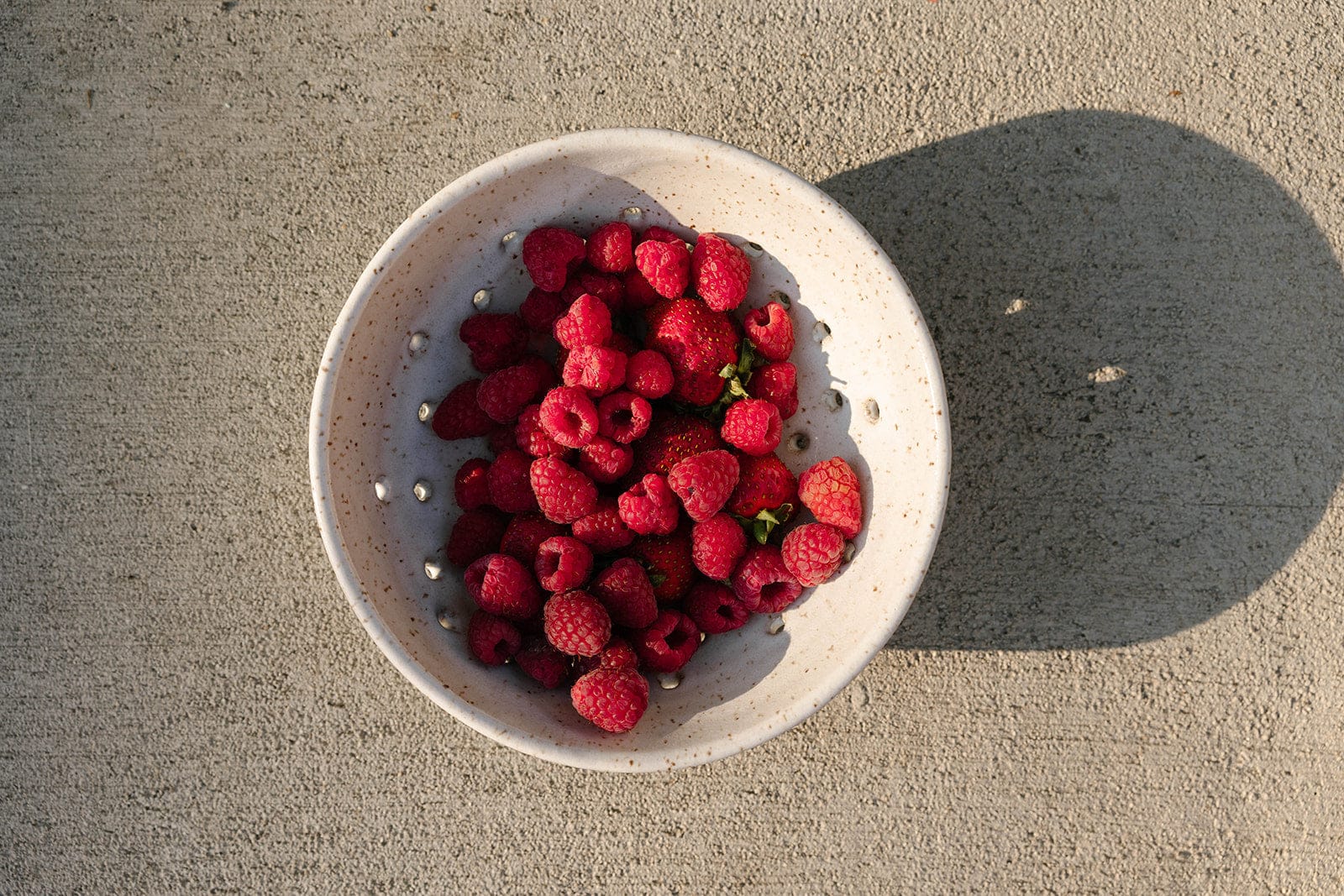 White ceramic colander filled with raspberries on a concrete surface