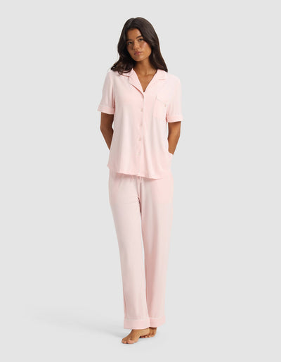 Front view of a model wearing pale pink pajamas with a short-sleeve collared button-front top and matching full-length pants, standing with her hands behind her back.