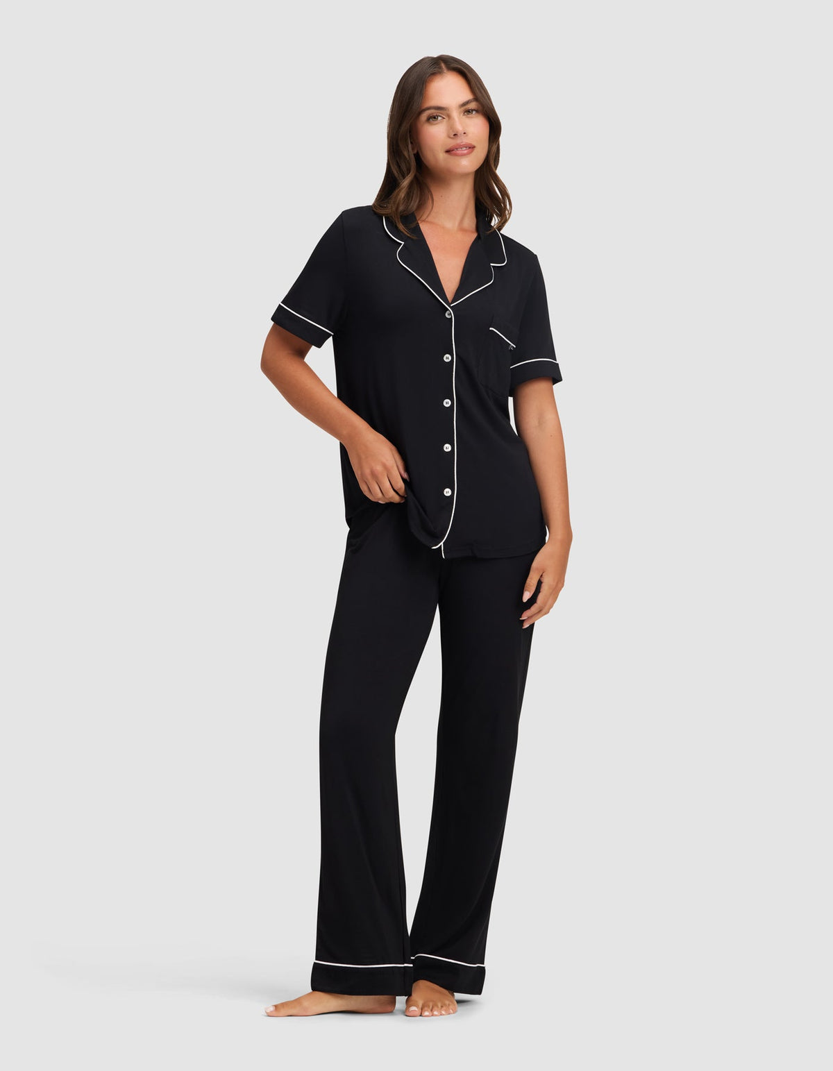 Front view of a model wearing black pajamas with a short-sleeve button-front top, notched collar, chest pocket, and relaxed pants, finished with contrasting white piping.