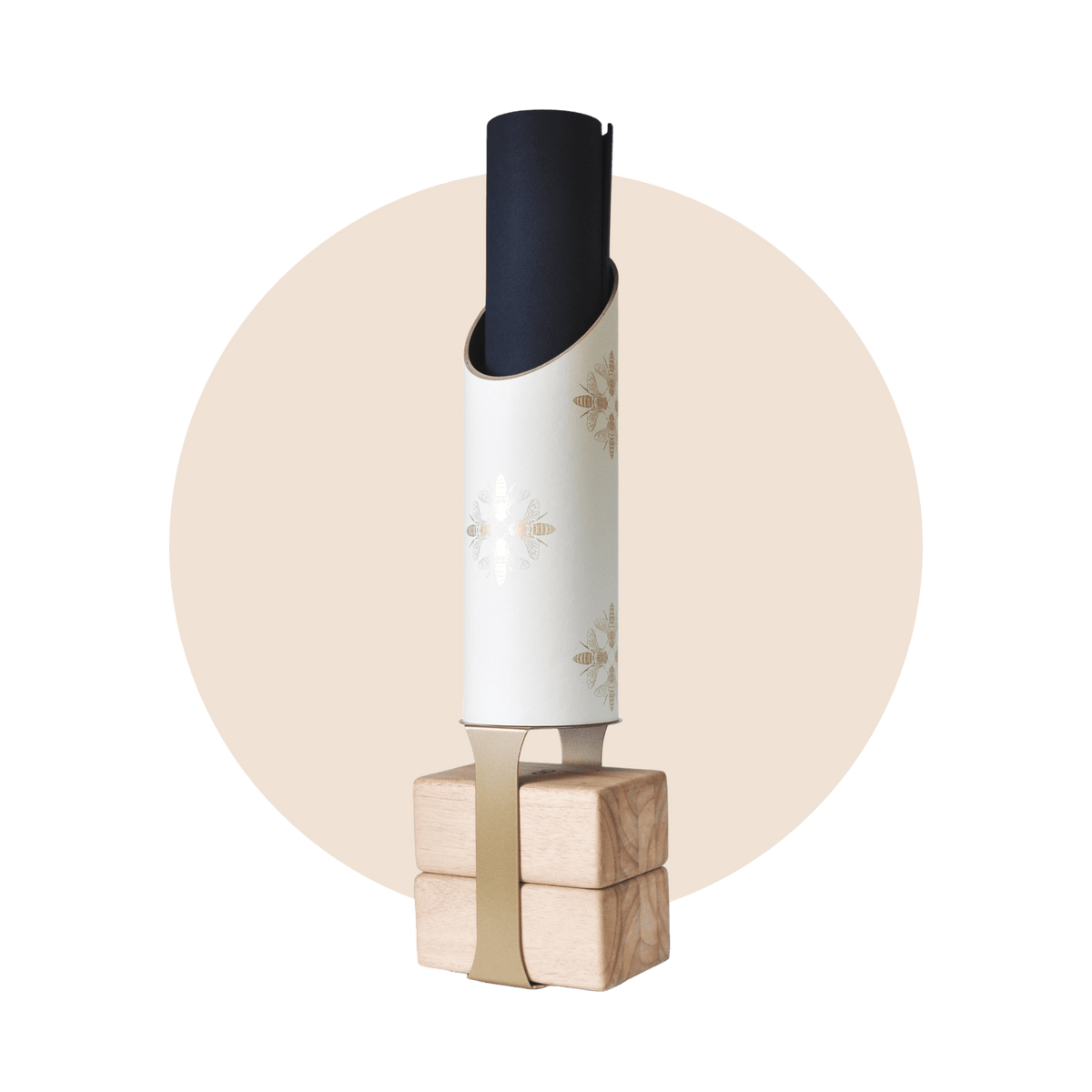 White cylindrical yoga storage tube decorated with delicate gold bee illustrations, holding a rolled black mat on four natural wood blocks with a tan support strap.