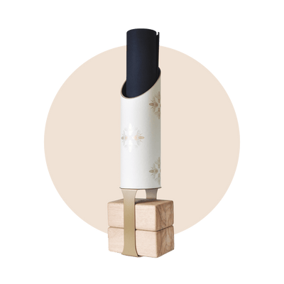 White cylindrical yoga storage tube decorated with delicate gold bee illustrations, holding a rolled black mat on four natural wood blocks with a tan support strap.