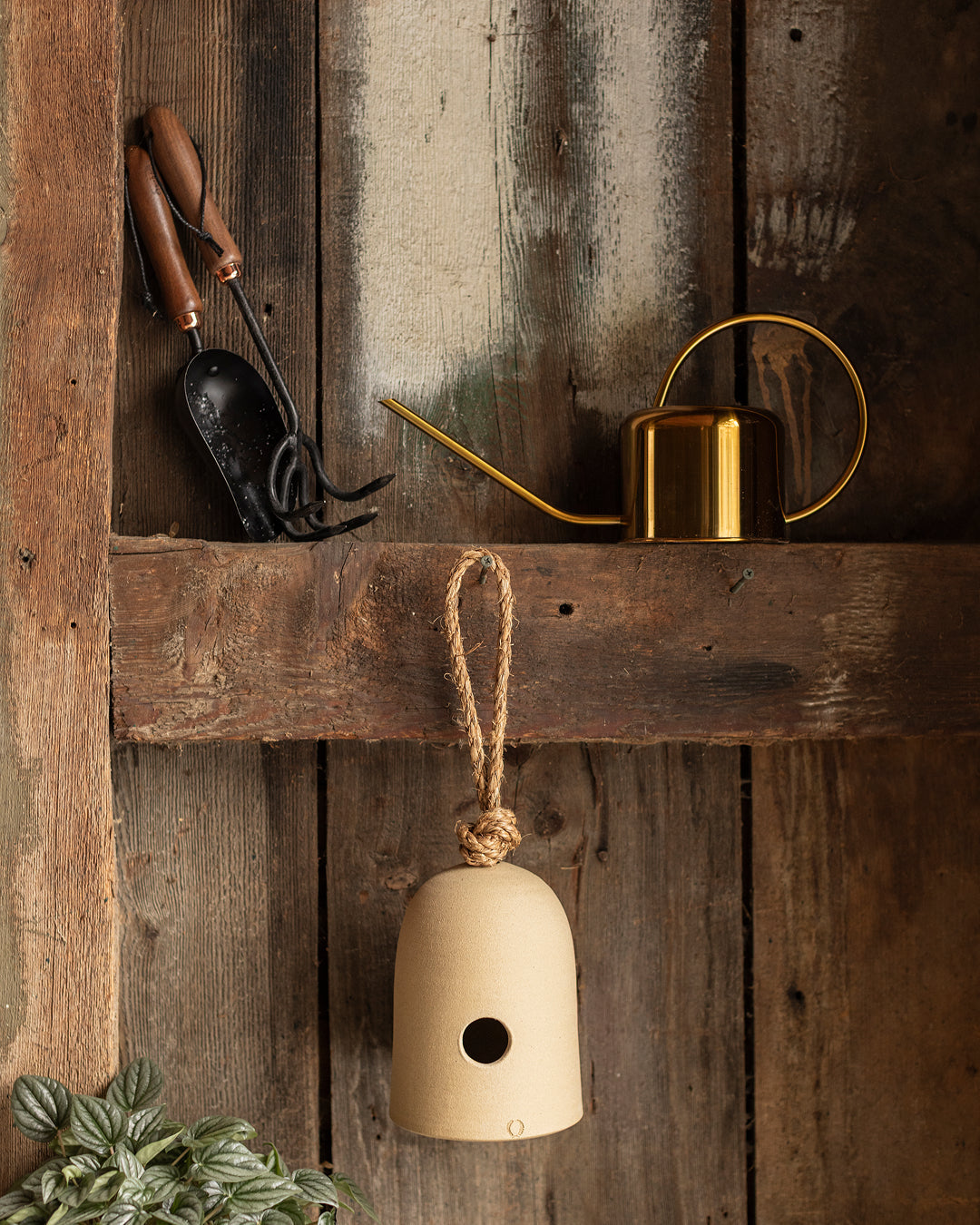 Fieldstone bird shelter hanging from a rough wooden beam in a rustic shed beside gardening shears, a brass watering can, and evergreen branches.