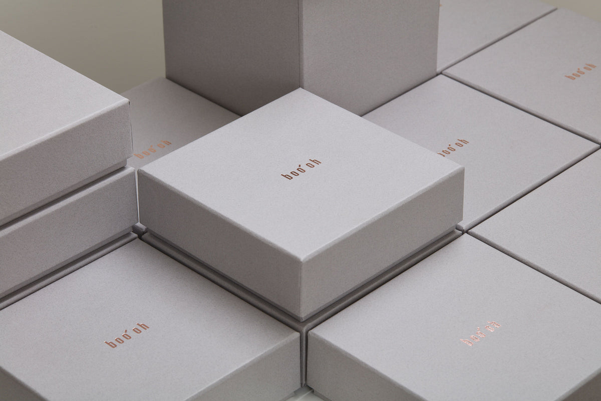 Stack of minimalist pale gray Boo Oh product boxes with small centered metallic lettering.