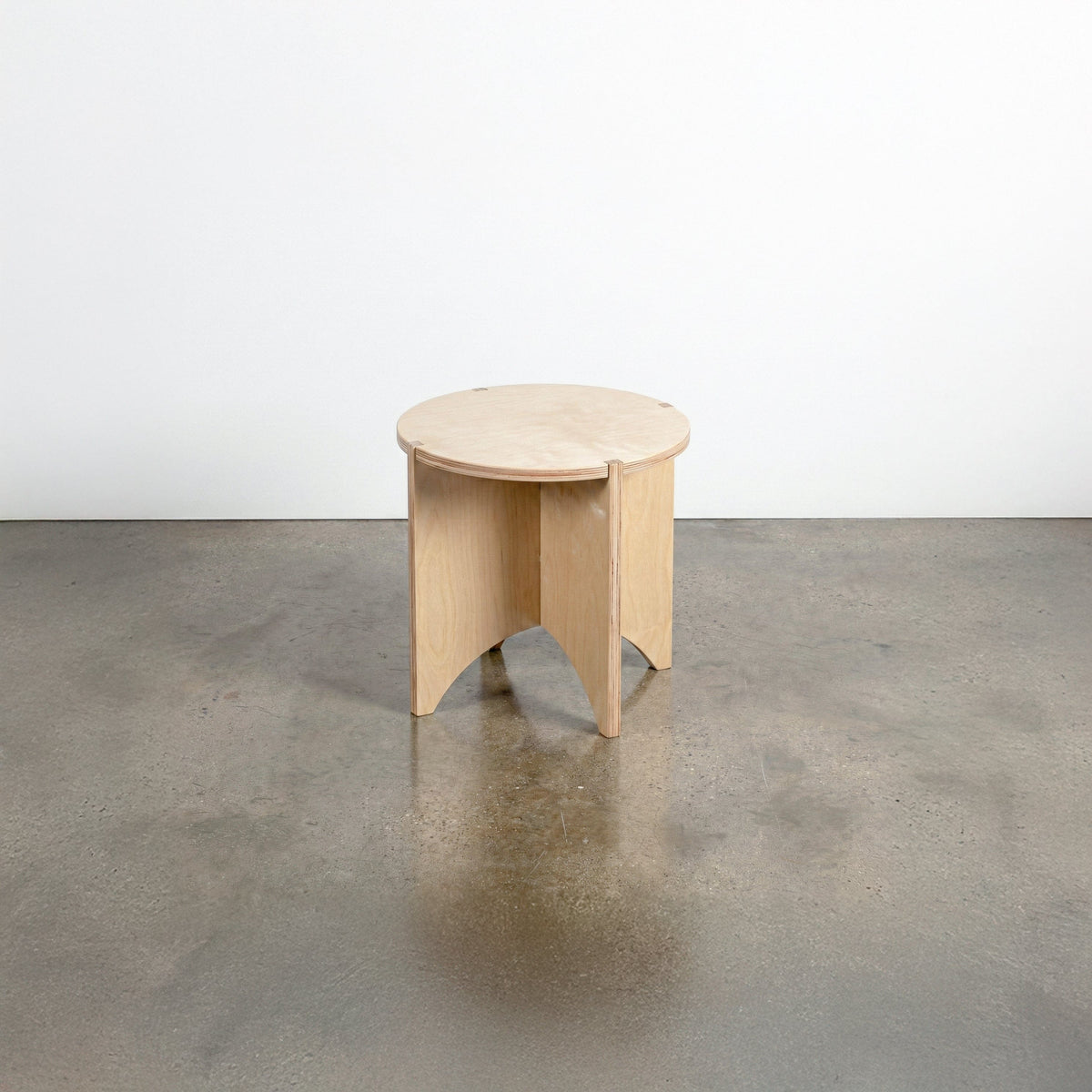 Studio Furnis YOYO natural wood end table with a round top and sculptural intersecting panel legs, displayed on a polished concrete floor
