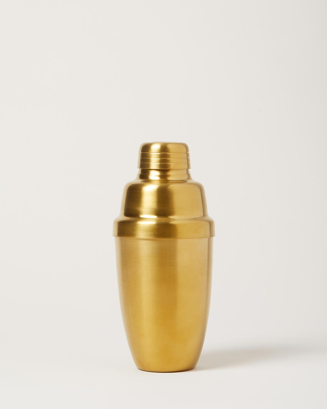 Brushed-gold cocktail shaker with a rounded body, fitted lid, and removable cap against a pale studio background.
