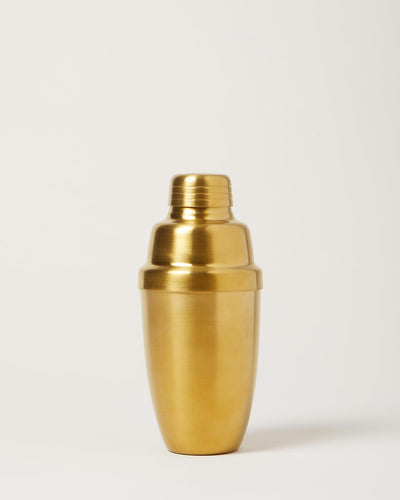 Brushed-gold cocktail shaker with a rounded body, fitted lid, and removable cap against a pale studio background.
